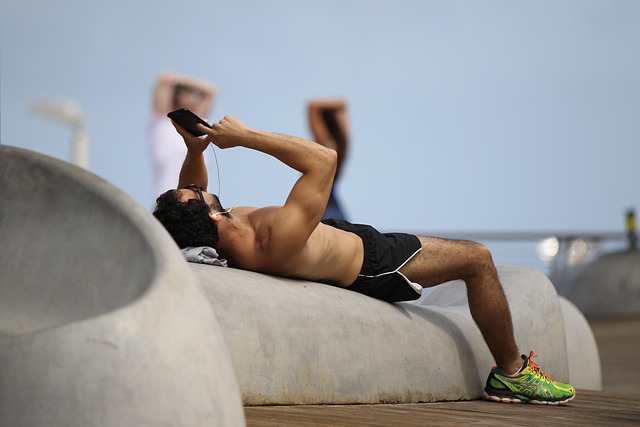 Un uomo in abbigliamento sportivo è sdraiato su una panchina di cemento all’aperto, con le cuffie nelle orecchie e uno smartphone in mano — forse sta controllando la sua app preferita per la corsa. Sullo sfondo si vedono due figure sfocate che fanno stretching.