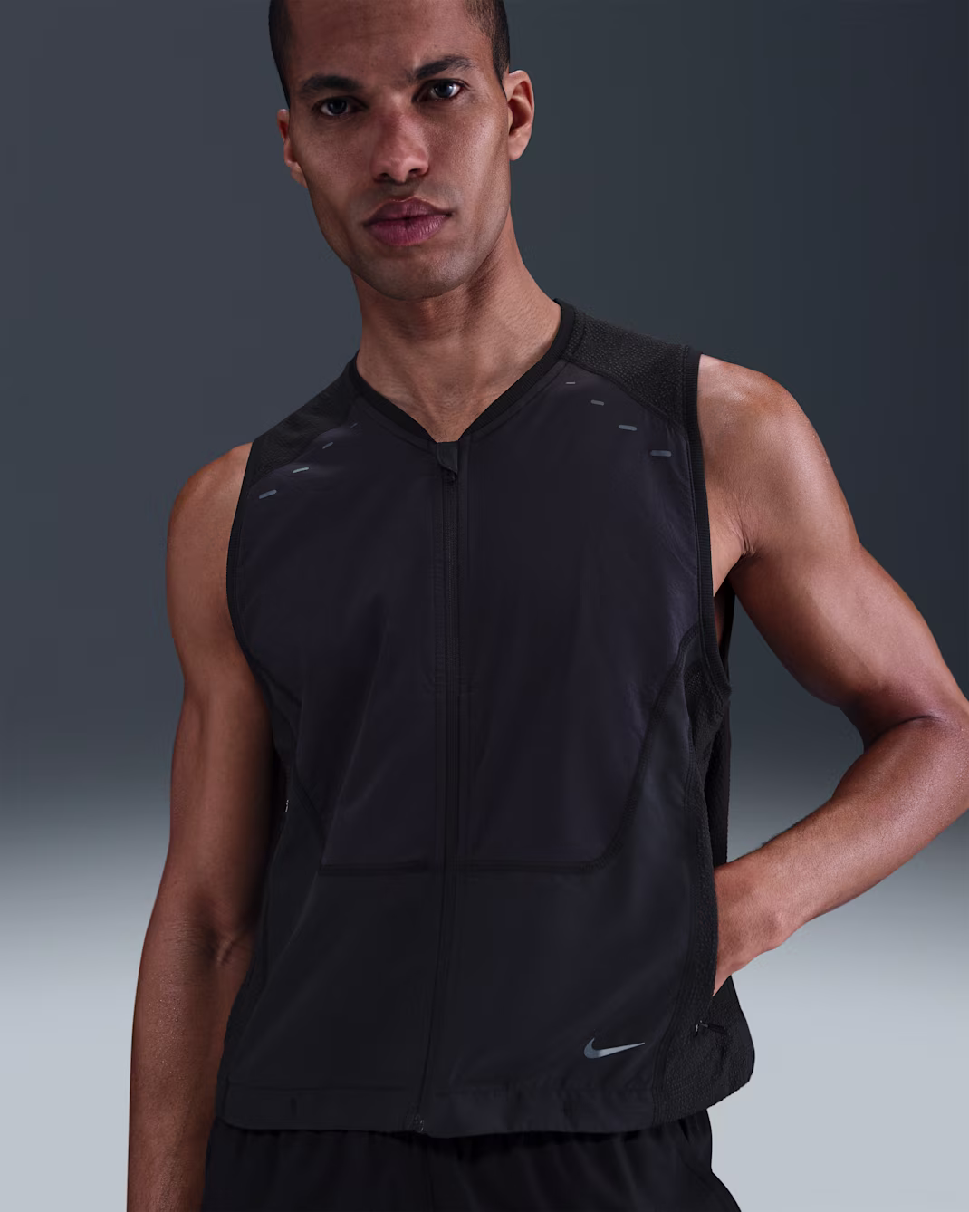 Una persona che indossa un gilet nero senza maniche con zip e un piccolo logo Nike si trova su uno sfondo a tinta unita sfumato.