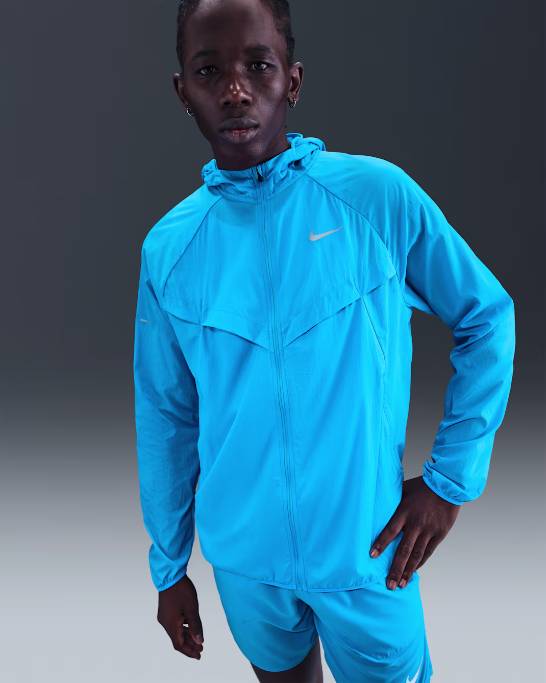 Una persona che indossa una giacca con cappuccio Nike blu brillante e pantaloncini abbinati si trova su uno sfondo neutro sfumato.