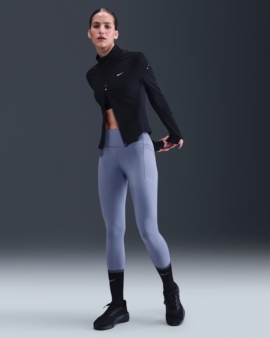 Una persona in piedi su uno sfondo semplice indossa una giacca nera, leggings grigi, calze nere e scarpe da ginnastica nere, con le braccia parzialmente distese dietro la schiena.