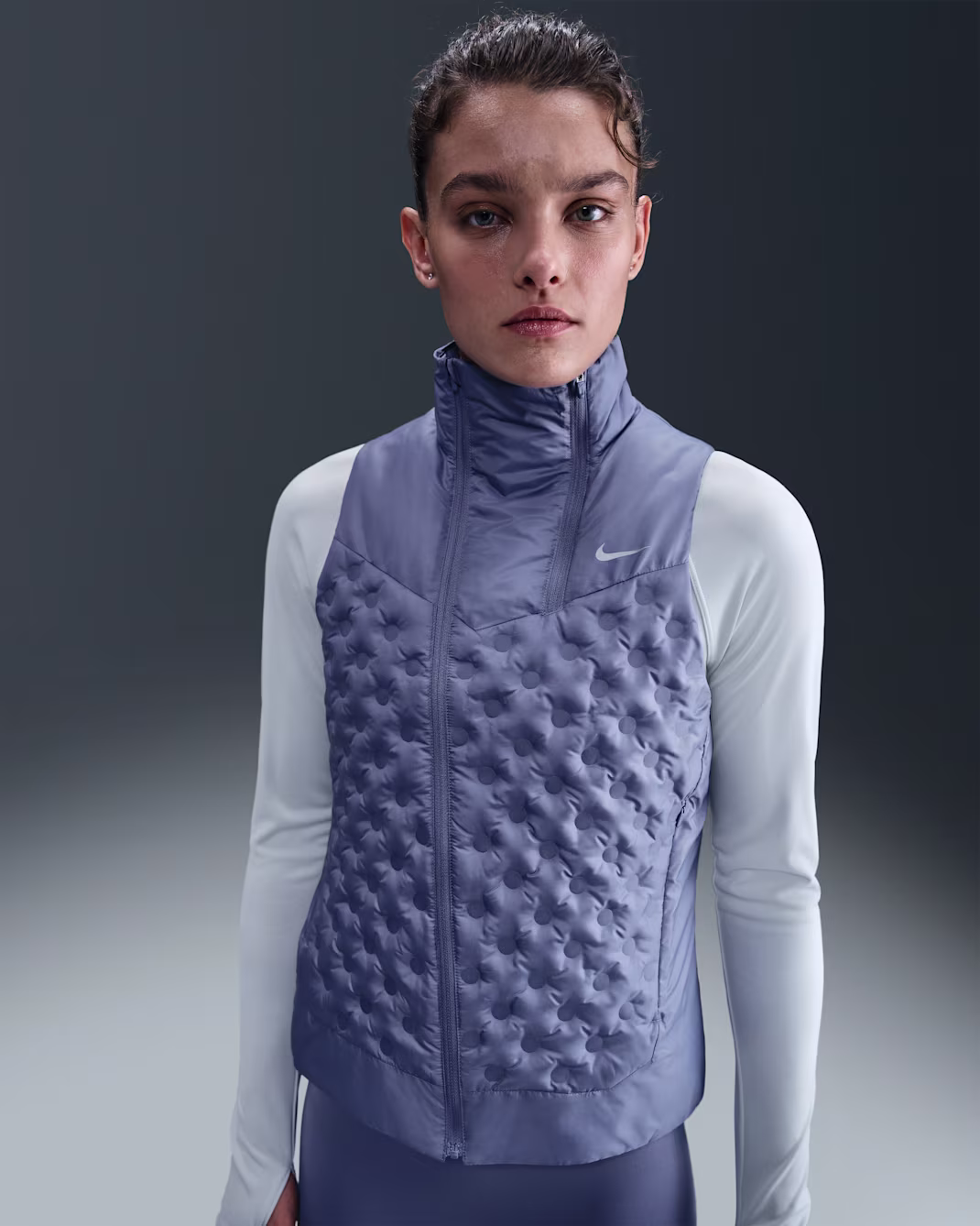 Una donna che indossa un gilet Nike trapuntato viola chiaro su un top atletico bianco a maniche lunghe è in piedi davanti a uno sfondo chiaro e scuro.